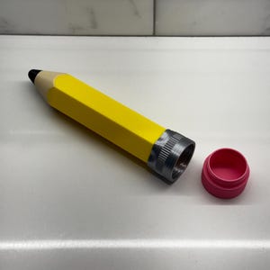 May include: A yellow pencil-shaped container with a silver lid and a pink lid. The container is open and the pink lid is off.