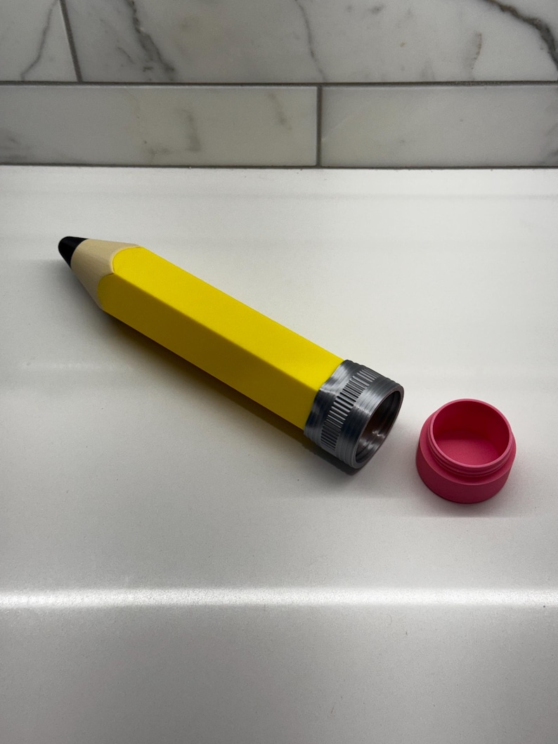 May include: A yellow pencil-shaped container with a silver lid and a pink lid. The container is open and the pink lid is off.