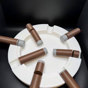 K&ouml;nnte beinhalten: Ein wei&szlig;er Aschenbecher mit kreisf&ouml;rmigem Design, der mehrere braune, zigarrenf&ouml;rmige Objekte mit grauen Spitzen aufweist. Die Objekte sind um den Aschenbecher herum angeordnet und bilden eine ansprechende Anordnung vor schwarzem Hintergrund.