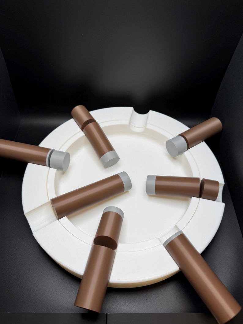 K&ouml;nnte beinhalten: Ein wei&szlig;er Aschenbecher mit kreisf&ouml;rmigem Design, der mehrere braune, zigarrenf&ouml;rmige Objekte mit grauen Spitzen aufweist. Die Objekte sind um den Aschenbecher herum angeordnet und bilden eine ansprechende Anordnung vor schwarzem Hintergrund.