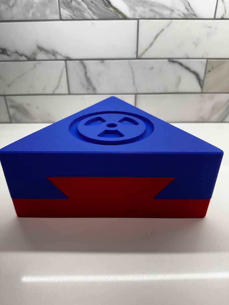 May include: A blue triangular box with a red bottom. The top of the box has a raised, circular design with a symbol that resembles a radiation symbol.