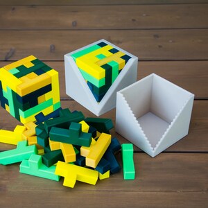 Puede incluir: Un rompecabezas geom&eacute;trico verde, amarillo y negro con una caja triangular blanca. El rompecabezas est&aacute; formado por peque&ntilde;os bloques que se pueden organizar de diferentes maneras. La caja est&aacute; abierta y el rompecabezas est&aacute; parcialmente ensamblado en su interior.