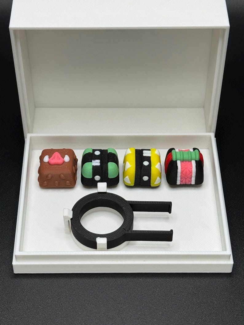May include: A white box with a black ring and four colorful keycaps. The keycaps are decorated with geometric patterns and a cartoon face.