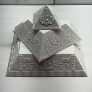 Puede incluir: Un conjunto de pir&aacute;mides grises impresas en 3D con intrincadas tallas y s&iacute;mbolos jerogl&iacute;ficos. El conjunto incluye tres pir&aacute;mides de diferentes tama&ntilde;os, apiladas para crear un efecto de capas. Las pir&aacute;mides presentan dise&ntilde;os intrincados y motivos del antiguo Egipto.
