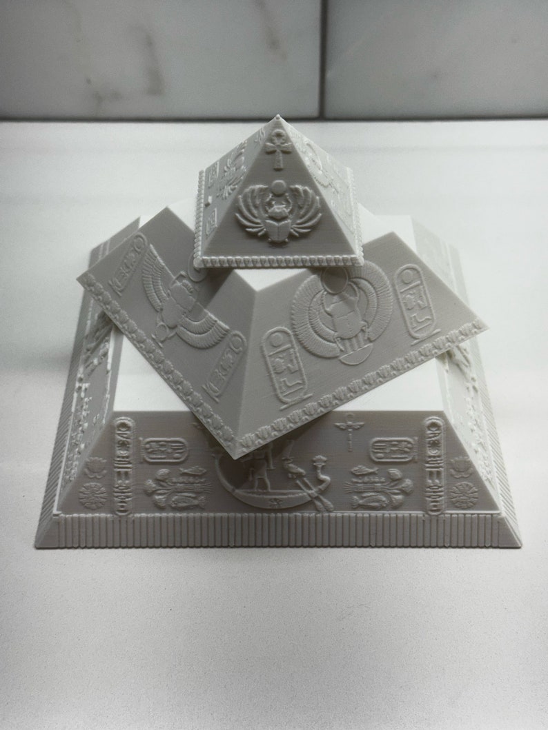 Puede incluir: Un conjunto de pir&aacute;mides grises impresas en 3D con intrincadas tallas y s&iacute;mbolos jerogl&iacute;ficos. El conjunto incluye tres pir&aacute;mides de diferentes tama&ntilde;os, apiladas para crear un efecto de capas. Las pir&aacute;mides presentan dise&ntilde;os intrincados y motivos del antiguo Egipto.