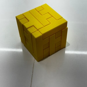 Puede incluir: Un cubo de rompecabezas 3D amarillo con un dise&ntilde;o complejo de entrelazado. El cubo est&aacute; hecho de pl&aacute;stico y tiene un acabado liso y mate.