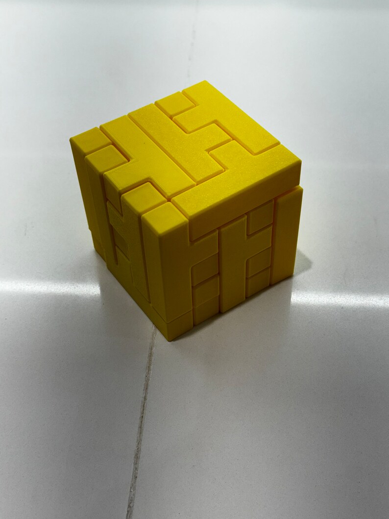 Puede incluir: Un cubo de rompecabezas 3D amarillo con un dise&ntilde;o complejo de entrelazado. El cubo est&aacute; hecho de pl&aacute;stico y tiene un acabado liso y mate.