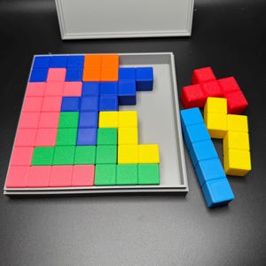 May include: A wooden puzzle game with a gray base and a lid. The puzzle pieces are made of colorful blocks in red, blue, green, yellow, and orange. The pieces are arranged in a pattern on the base.