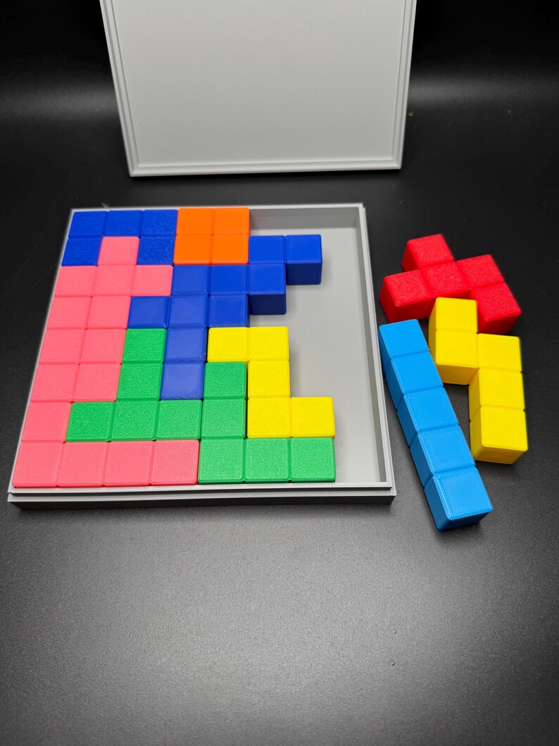 May include: A wooden puzzle game with a gray base and a lid. The puzzle pieces are made of colorful blocks in red, blue, green, yellow, and orange. The pieces are arranged in a pattern on the base.