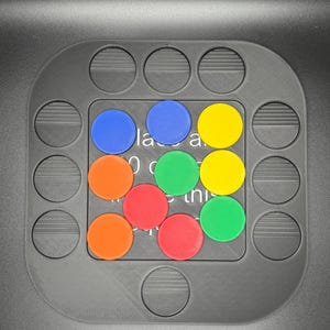 K&ouml;nnte beinhalten: Ein graues Puzzlespielbrett mit kreisf&ouml;rmigen Aussparungen und einem zentralen Raster. Das Raster enth&auml;lt farbige kreisf&ouml;rmige Teile in Blau, Orange, Rot, Gr&uuml;n und Gelb. Text ist auf dem Brett sichtbar. Ein lustiges und ansprechendes Spiel.