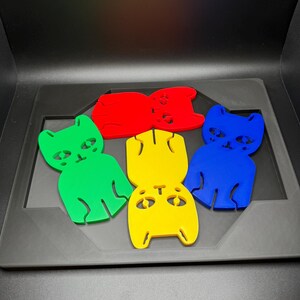 May include: Five colorful cat-shaped cutouts in red, green, yellow, and blue are arranged on a gray tray. Each cutout has a simple design with eyes, a nose, and a mouth. The background is black.