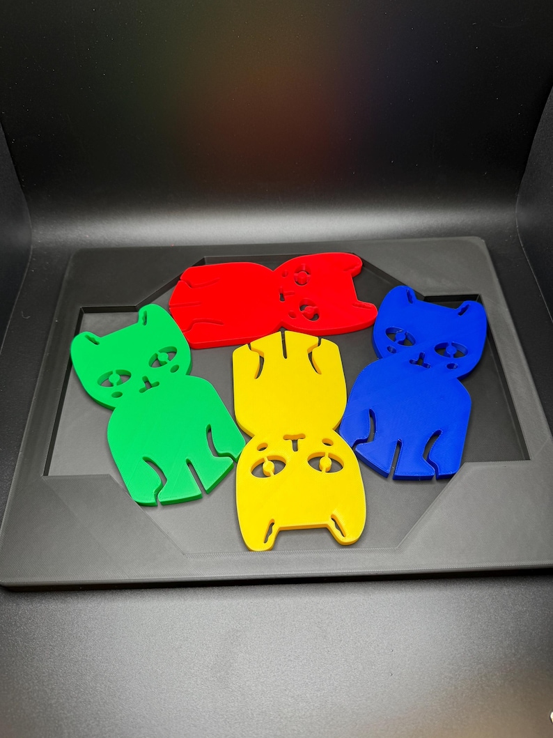May include: Five colorful cat-shaped cutouts in red, green, yellow, and blue are arranged on a gray tray. Each cutout has a simple design with eyes, a nose, and a mouth. The background is black.