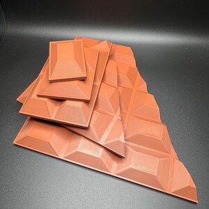 Op de afbeelding: Een set van zes terracottakleurige, geometrisch gepatroneerde, 3D-geprinte tegels. De tegels zijn gerangschikt in een piramidevorm, met de kleinste tegel bovenaan en de grootste tegel onderaan.