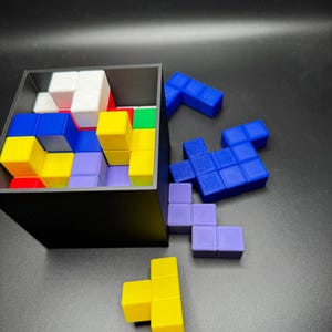 May include: A black box filled with colorful, geometric puzzle pieces. The pieces are various shapes and colors, including blue, yellow, red, green, white, and purple. Some pieces are outside the box, scattered on a dark surface.