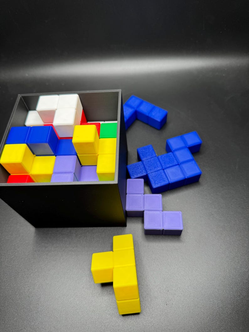 May include: A black box filled with colorful, geometric puzzle pieces. The pieces are various shapes and colors, including blue, yellow, red, green, white, and purple. Some pieces are outside the box, scattered on a dark surface.