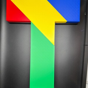 May include: A three-dimensional letter T constructed from geometric segments. The top horizontal bar features a red section on the left, a central yellow triangle, and a blue section on the right. A vertical green rectangular base extends downward.