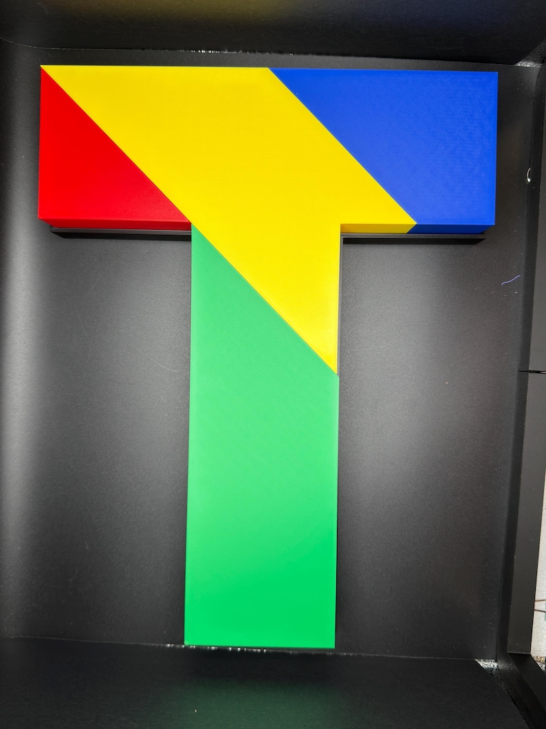 May include: A three-dimensional letter T constructed from geometric segments. The top horizontal bar features a red section on the left, a central yellow triangle, and a blue section on the right. A vertical green rectangular base extends downward.