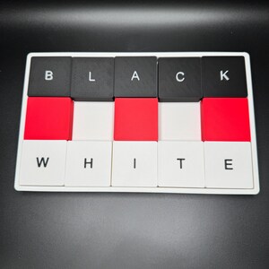 May include: A rectangular game board with black, red, and white tiles. The top row spells "BLACK" and the bottom row spells "WHITE". The tiles are arranged in a grid pattern, with the letters in black on a white background.