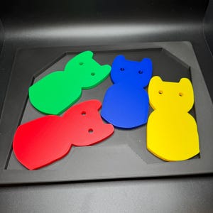 May include: Four colorful cat-shaped cutouts in red, green, blue, and yellow. Each cutout has two small holes and is displayed on a black tray.