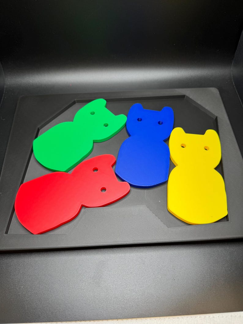 May include: Four colorful cat-shaped cutouts in red, green, blue, and yellow. Each cutout has two small holes and is displayed on a black tray.