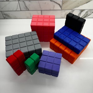 May include: A set of nine colorful, square-shaped blocks with a textured surface. The blocks are made of a soft, pliable material and are arranged in a pyramid shape. The blocks are in various colors, including red, orange, yellow, green, blue, purple, and gray.