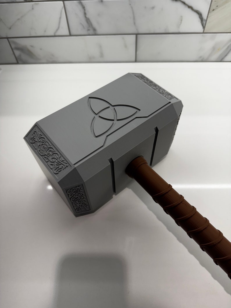 Peut inclure: Un marteau en plastique gris avec un manche brun. Le marteau a un motif en relief sur la t&ecirc;te, y compris un symbole &agrave; trois pointes.