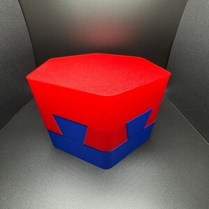May include: A red and blue geometric puzzle box with a hexagonal shape. The box is made of plastic and has a smooth, glossy finish.
