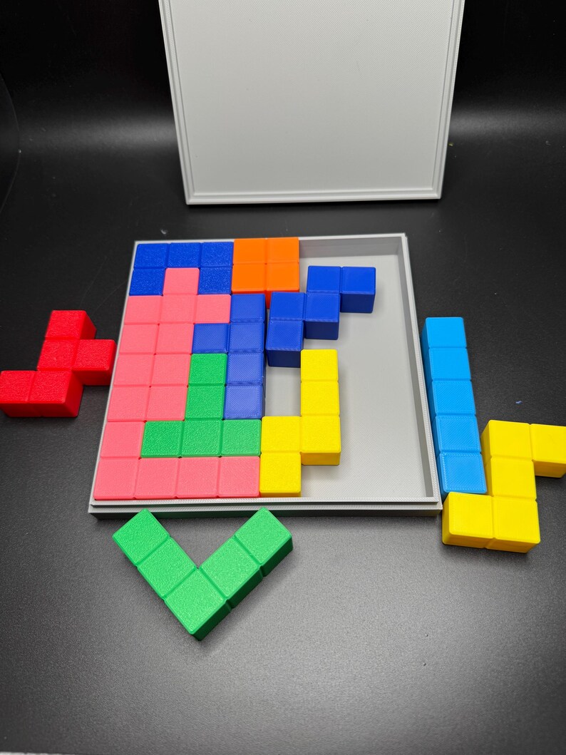 May include: A wooden puzzle game with colorful blocks in red, blue, green, yellow, and orange. The blocks are arranged in a grid pattern on a gray tray. The game is in a partially open box.