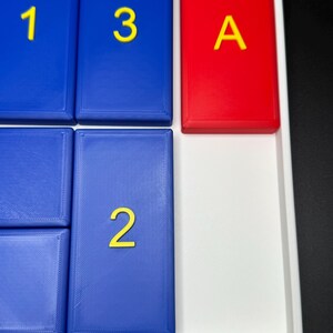 May include: A white tray with blue and red tiles. The blue tiles have the numbers 1, 2, and 3 printed in yellow. The red tile has the letter A printed in yellow.