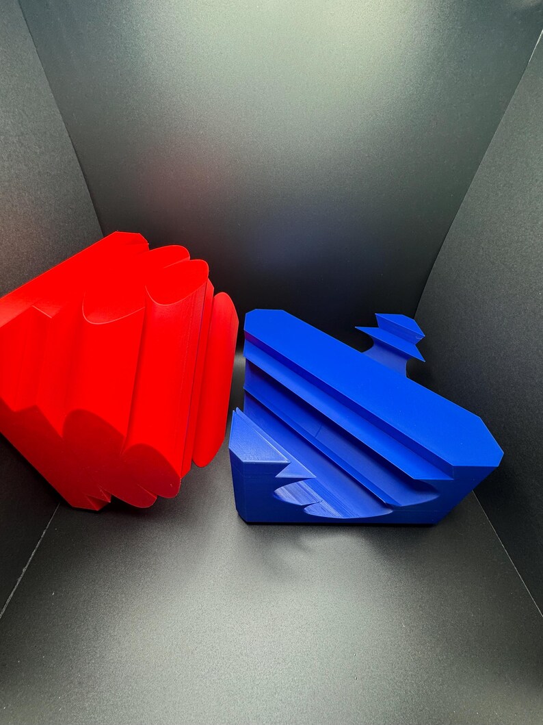 May include: Two geometric shapes, one red and one blue, made of a smooth, solid material. The red shape is a multi-sided, rounded object. The blue shape is a flat, multi-sided object with a curved edge.
