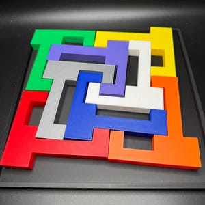 Op de afbeelding: Een kleurrijke puzzel met zeven stukken, elk in de vorm van een T, past samen om een vierkant te vormen. De stukken zijn rood, oranje, geel, groen, blauw, paars en grijs.