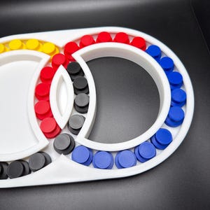 May include: A white plastic frame featuring a series of circular slots filled with small, cylindrical pegs in red, blue, yellow, and black. The structure consists of two interconnected circular sections with a central almond-shaped divider.