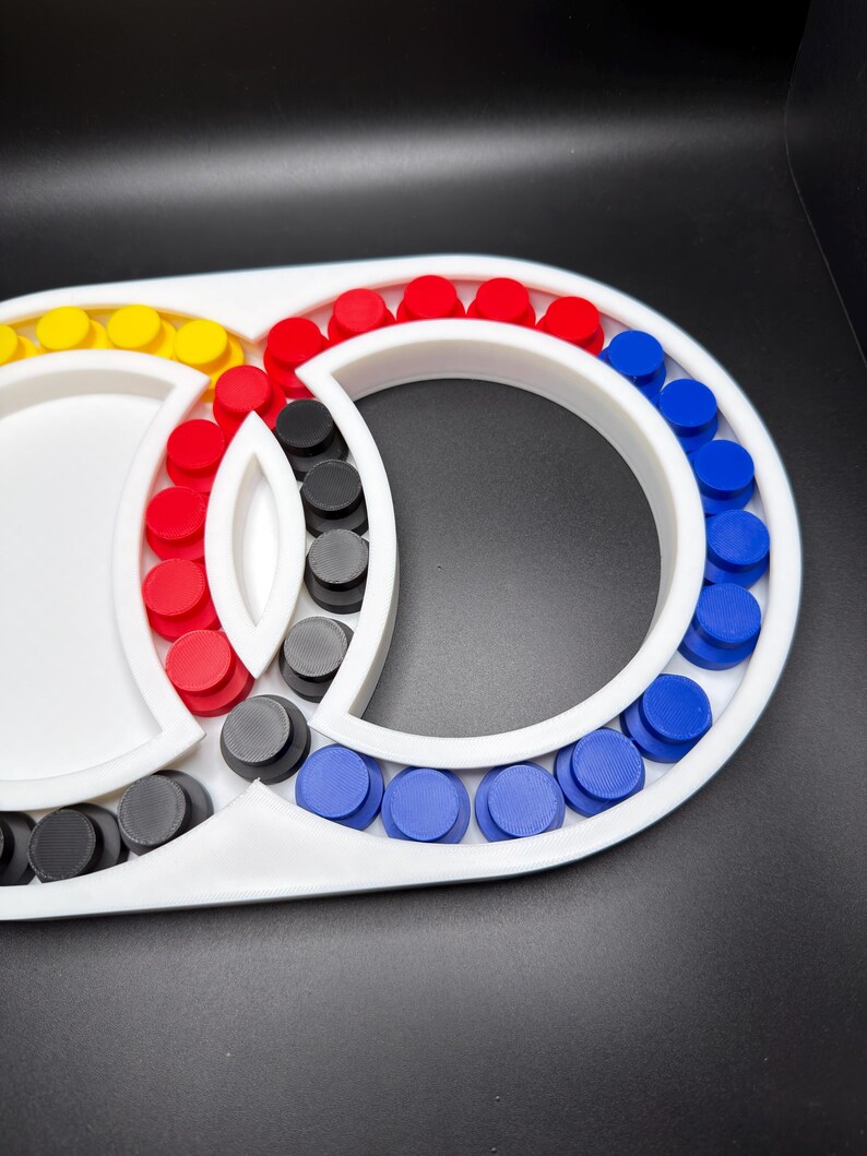 May include: A white plastic frame featuring a series of circular slots filled with small, cylindrical pegs in red, blue, yellow, and black. The structure consists of two interconnected circular sections with a central almond-shaped divider.