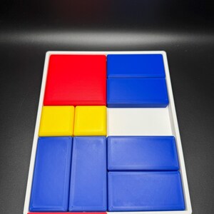 Op de afbeelding: Een wit dienblad met een geometrisch ontwerp in rood, geel en blauw. Het dienblad is verdeeld in negen secties, elk met een blok in een andere kleur. De blokken zijn gerangschikt in een patroon dat doet denken aan een schilderij van Mondrian.