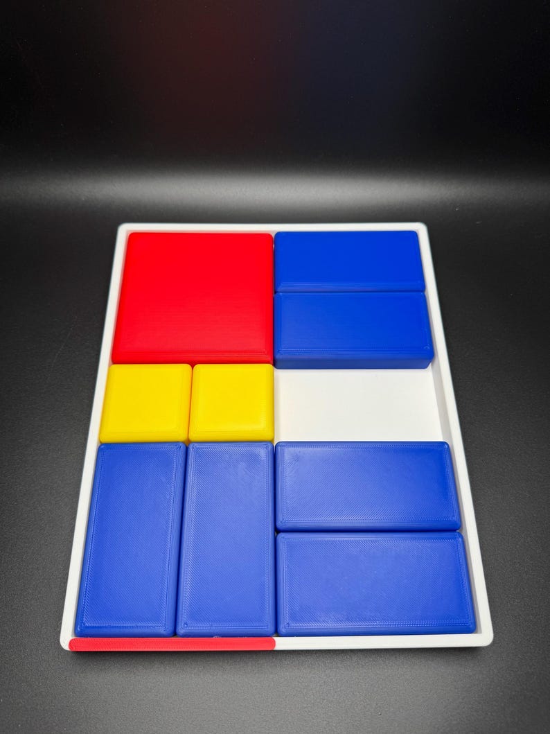 Op de afbeelding: Een wit dienblad met een geometrisch ontwerp in rood, geel en blauw. Het dienblad is verdeeld in negen secties, elk met een blok in een andere kleur. De blokken zijn gerangschikt in een patroon dat doet denken aan een schilderij van Mondrian.