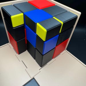 May include: A wooden box with a lid that opens to reveal a 3D puzzle made of small cubes. The cubes are arranged in a pattern of black, red, blue, and yellow.