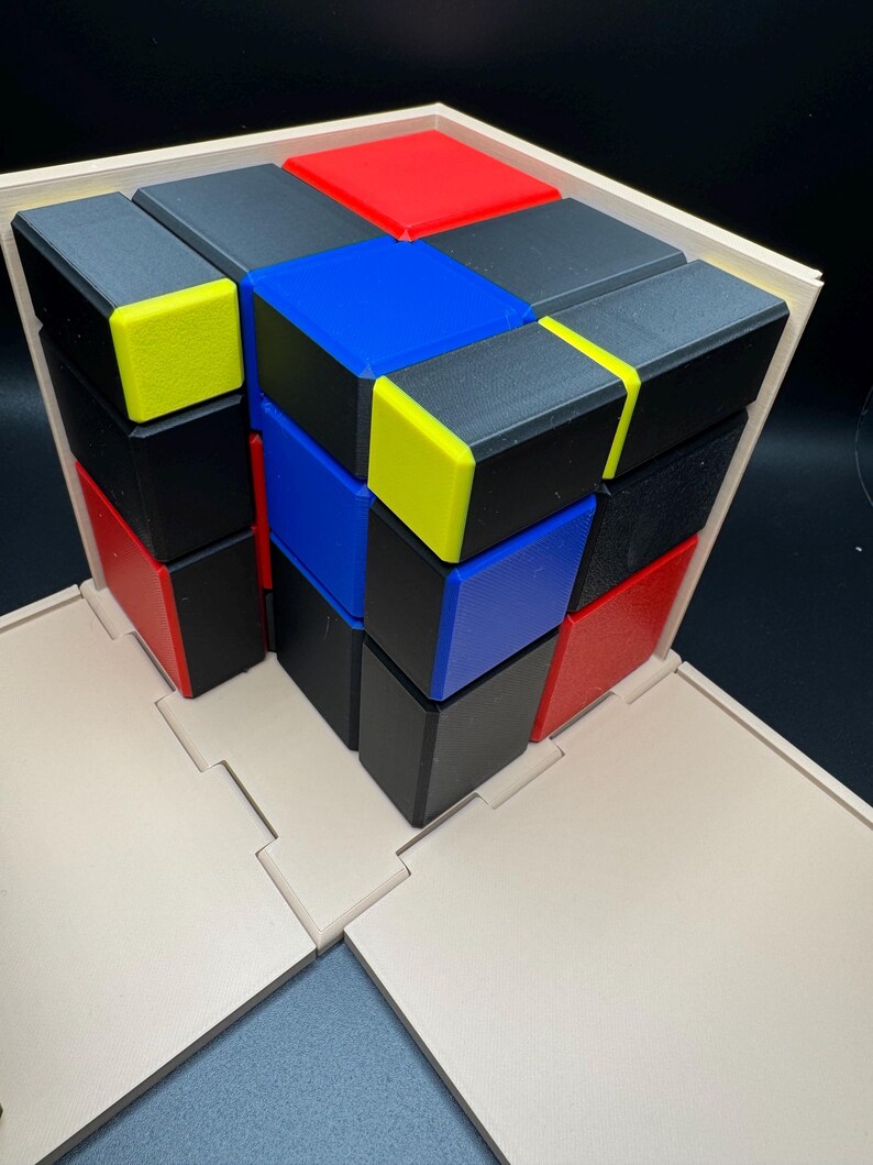 May include: A wooden box with a lid that opens to reveal a 3D puzzle made of small cubes. The cubes are arranged in a pattern of black, red, blue, and yellow.