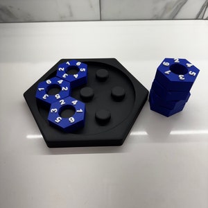 May include: A blue hexagonal puzzle with numbered pieces. The puzzle is made up of six pieces, each with a different number on it. The pieces are arranged in a circle, with the numbers facing outwards. The puzzle is on a black hexagonal base.