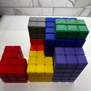 May include: A set of colorful building blocks made of plastic. The blocks are arranged in a tower with a red block on the bottom, a yellow block in the middle, and a blue block on top. There is a green block on top of the blue block and a gray block on top of the yellow block.