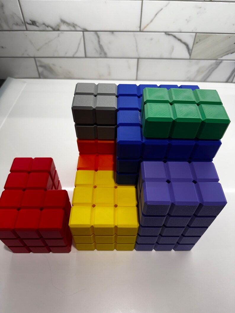 May include: A set of colorful building blocks made of plastic. The blocks are arranged in a tower with a red block on the bottom, a yellow block in the middle, and a blue block on top. There is a green block on top of the blue block and a gray block on top of the yellow block.