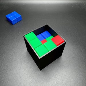 May include: A black square container filled with colorful, square puzzle pieces. The pieces are red, green, and blue. A separate blue puzzle piece sits to the side. The puzzle is on a gray surface.