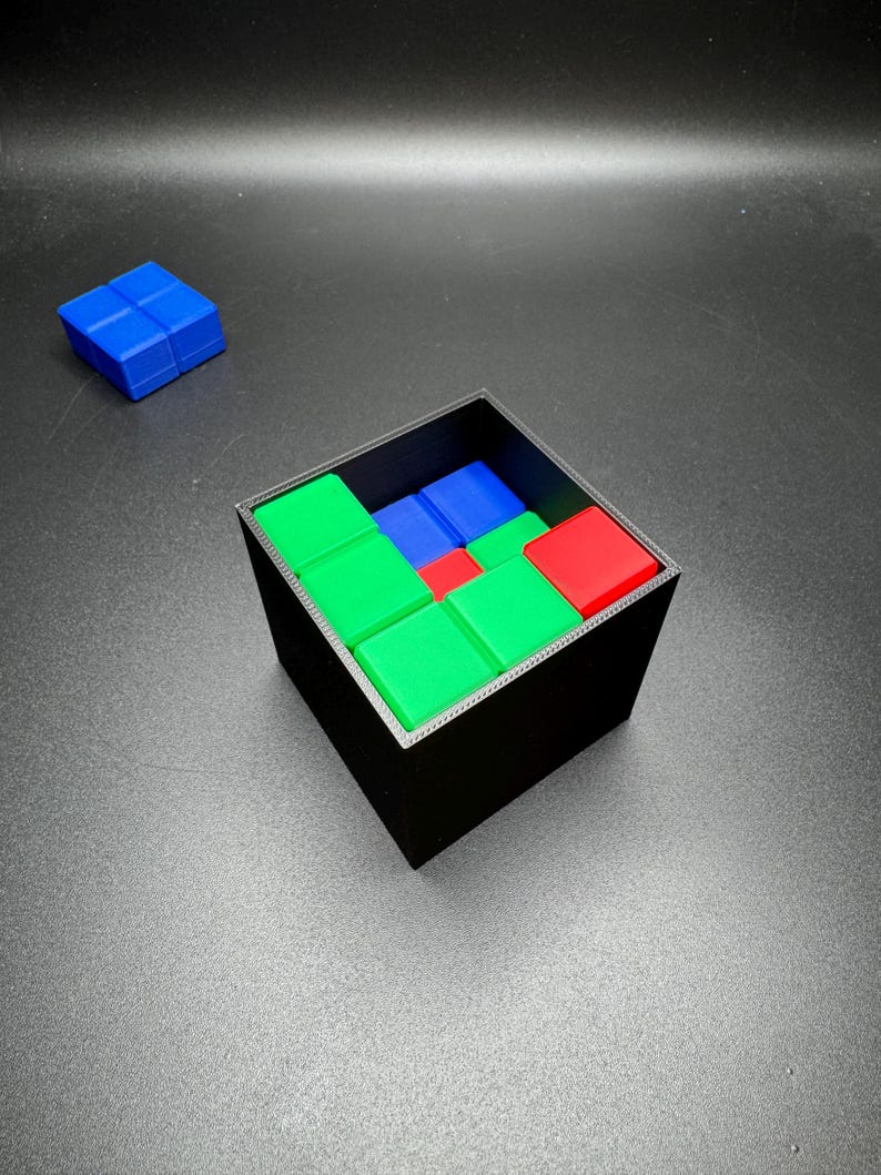 May include: A black square container filled with colorful, square puzzle pieces. The pieces are red, green, and blue. A separate blue puzzle piece sits to the side. The puzzle is on a gray surface.