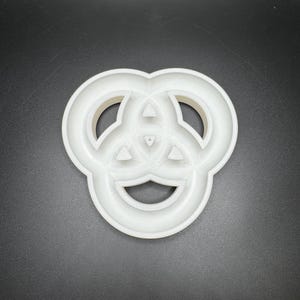 May include: A white plastic cookie cutter in the shape of a Celtic knot. The cutter has a three-lobed design with rounded edges and a central triangular detail. The object is set against a dark background.