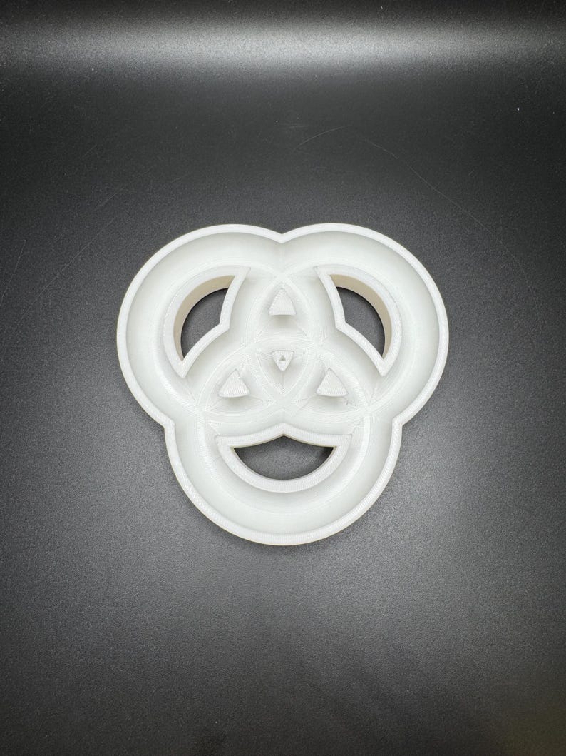 May include: A white plastic cookie cutter in the shape of a Celtic knot. The cutter has a three-lobed design with rounded edges and a central triangular detail. The object is set against a dark background.