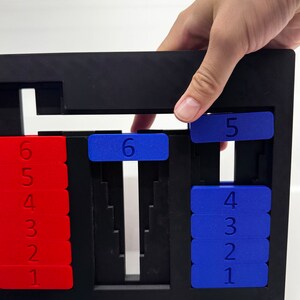 May include: A black plastic frame with red and blue number tiles. The tiles are arranged in columns, with the numbers 1 through 6 visible. A hand is reaching into the frame to move a blue tile with the number 5 on it.