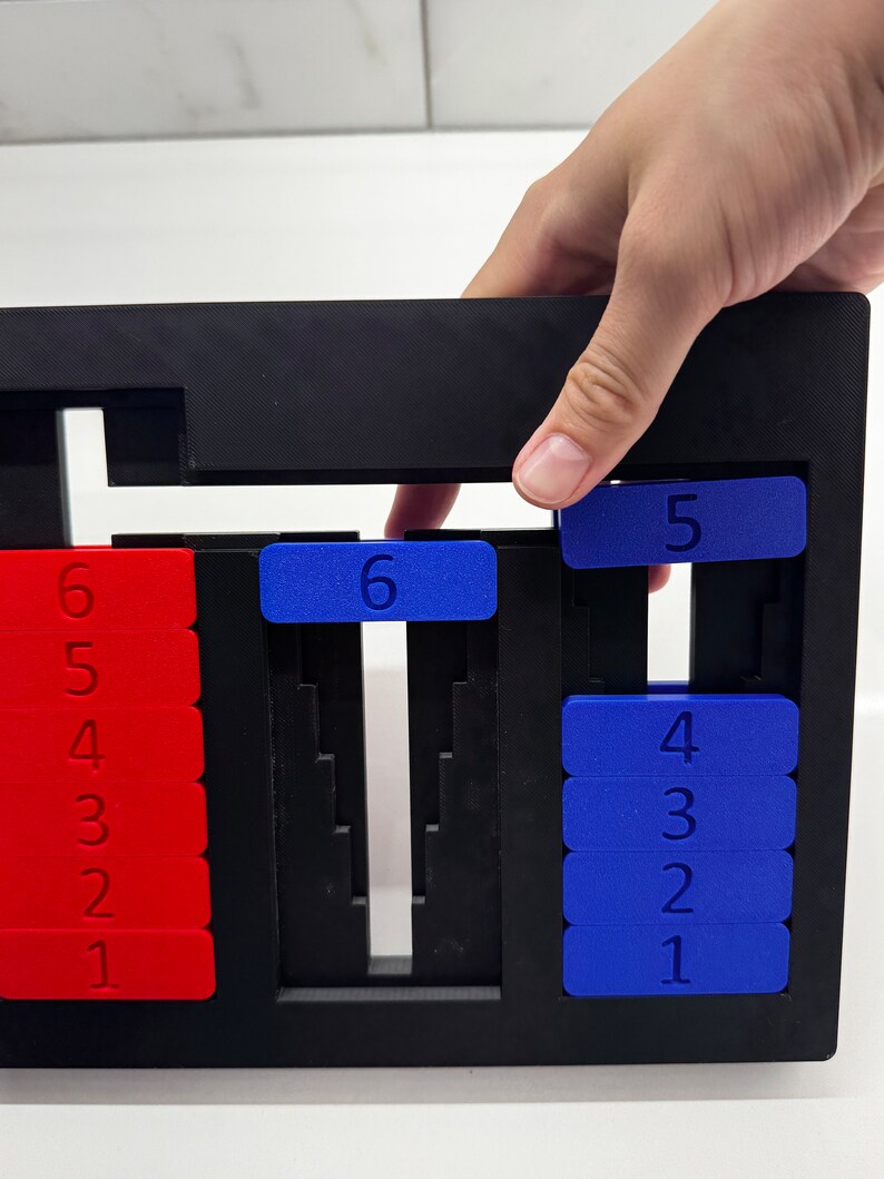 May include: A black plastic frame with red and blue number tiles. The tiles are arranged in columns, with the numbers 1 through 6 visible. A hand is reaching into the frame to move a blue tile with the number 5 on it.