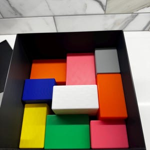 May include: A black box filled with colorful blocks. The blocks are arranged in a pattern and include red, orange, yellow, green, blue, white, pink, and gray.