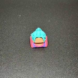 May include: A teal and purple keycap with a detailed design of a woman's face with a bun.