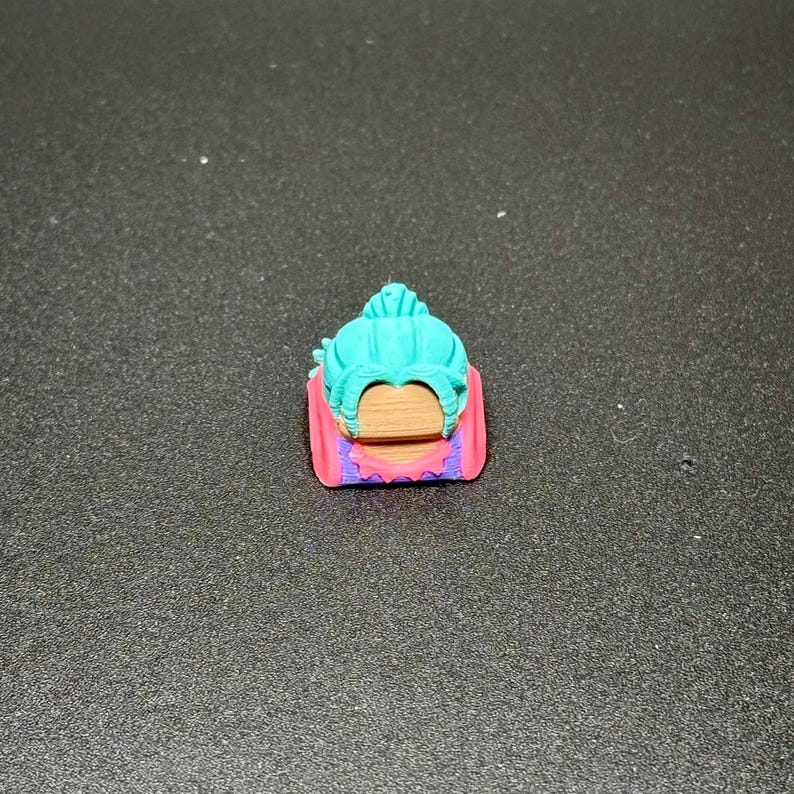 May include: A teal and purple keycap with a detailed design of a woman's face with a bun.