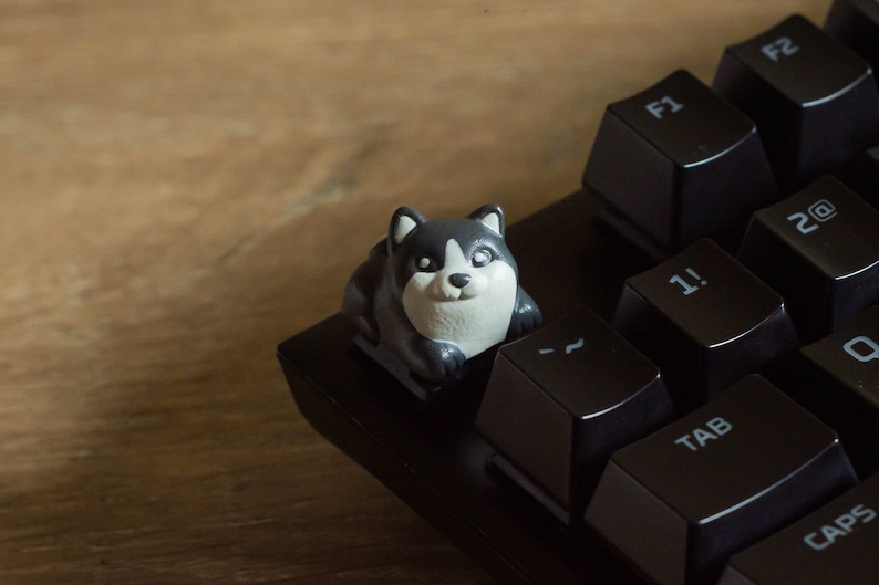 May include: A custom keycap shaped like a husky dog, in black, white, and gray, sits on a black keyboard. The keycap replaces a standard key. The keyboard keys are black with white lettering. The background is a wooden surface.
